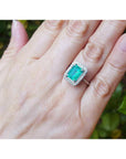 Emerald-cut bluish-green emerald ring with halo diamonds in 14K white gold on a woman's hand.