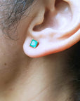 Close-up of a woman's ear wearing a square yellow gold emerald stud earring with hair and skin visible.