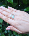 Hand holding two natural emerald stud earrings against a green leafy background.