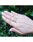 Hand holding two natural emerald stud earrings against a green leafy background.