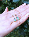 Hand displaying two emerald gemstone earrings with a green leafy background