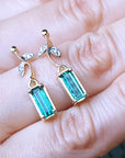 Gold earrings with green Muzo Colombian emeralds held between fingers on a light skin background