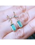 Gold earrings with green Muzo Colombian emeralds held between fingers on a light skin background