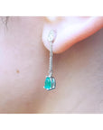 Pear-shaped Muzo Colombian emerald dangle earrings in 14K white gold with diamond accents on an ear.
