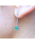 Pear-shaped Muzo Colombian emerald dangle earrings in 14K white gold with diamond accents on an ear.