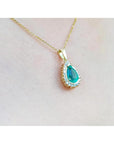 Teardrop emerald pendant with a diamond halo on a gold chain, worn on a woman's neck.