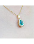 Teardrop emerald pendant with a diamond halo on a gold chain, worn on a woman's neck.