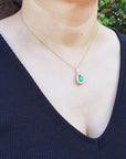 A woman wearing a gold necklace with a colombian emerald pendant against a blurred natural background