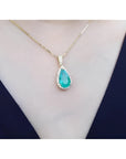 18K yellow gold teardrop emerald pendant with halo diamonds on a delicate chain, worn on a neck.