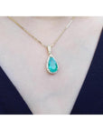 18K yellow gold teardrop emerald pendant with halo diamonds on a delicate chain, worn on a neck.