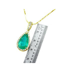 A natural colombian emerald teardrop pendant on a gold chain with a ruler for scale on a white background