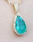 Teardrop-shaped emerald gemstone pendant with halo diamond accents on a light pink skin background