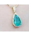 Teardrop-shaped emerald gemstone pendant with halo diamond accents on a light pink skin background