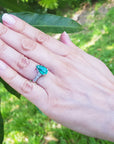 Hand wearing a teardrop emerald ring with greenery in the background.