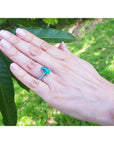 Hand wearing a teardrop emerald ring with greenery in the background.