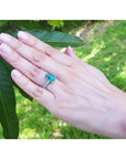 Pear-shaped deep bluish-green emerald ring with princess cut diamonds in 14K white gold on a hand.