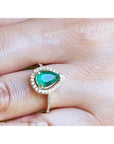 A pear cut emerald encircled by a halo diamond in yellow gold ring on a finger.