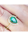A pear cut emerald in a halo diamond ring on a finger.