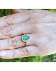 Hand wearing a gold ring with a pear cut emerald into a halo diamond against a blurred natural background.