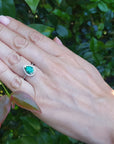 Hand wearing a teardrop emerald ring with a blurred green leafy background.