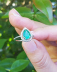Hand holding between finger a pear cut emerald ring against a blurred green leafy background.