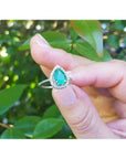 14K white gold teardrop emerald ring with diamond halo, held against a leafy background.