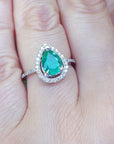 Ring with a natural pear shaped emerald on a person's finger against a blurred natural background.