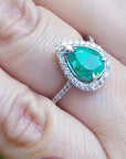 Emerald ring with diamond accents on a finger against a blurred natural background.