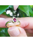 14K yellow gold teardrop natural ruby solitaire ring, held by fingers against green leaves.