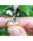 14K yellow gold teardrop natural ruby solitaire ring, held by fingers against green leaves.