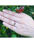 Woman's hand wearing a 14K yellow gold teardrop natural ruby solitaire ring, against green foliage.