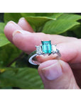 A natural three stone emerald ring for women held in a hand against a green leafy background.