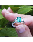 18K white gold three-stone emerald cut ring with a medium bluish-green emerald and side baguette diamonds.
