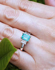 An emerald three stone emerald ring on a hand with green leaves in the background.