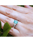 An emerald three stone emerald ring on a hand with green leaves in the background.