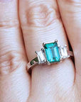 A three stone emerald and side straight baguettes diamond ring on a finger against a natural background.