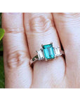 A three stone emerald and side straight baguettes diamond ring on a finger against a natural background.