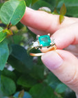 A Colombian emerald ring for women held between fingers against a green leafy background.