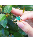 A Colombian emerald ring for women held between fingers against a green leafy background.