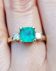 A three stone emerald engagement rings with a green gemstone on a finger.