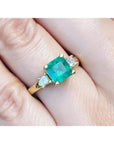 Emerald ring for women with gold setting and side princess cut natural diamonds on a finger.