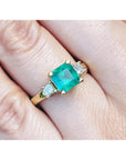 Emerald ring for women with gold setting and side princess cut natural diamonds on a finger.