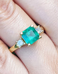 Emerald ring with gold setting and side diamonds on a finger.