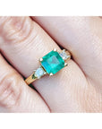 Emerald ring with gold setting and side diamonds on a finger.