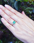 Hand wearing a natural emerald ring with side diamonds against a leafy background.