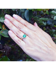 Hand wearing a natural emerald ring with side diamonds against a leafy background.