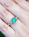 Emerald ring on a finger with a blurred green background.