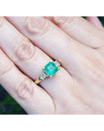 Emerald ring on a finger with a blurred green background.