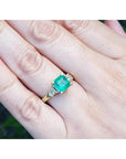 18K yellow gold three-stone ring with a square emerald and princess-cut side diamonds on a hand.