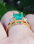 Gold ring with a natural emerald on a finger against a blurred green background.